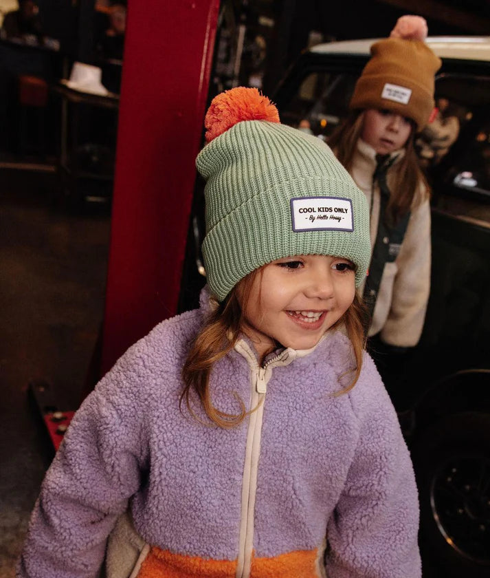 Menina com gorro Hello Hossy verde-menta e pompom laranja, vista lateral, num ambiente urbano.