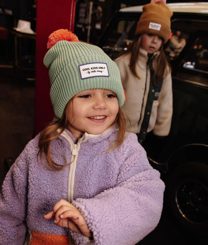 Menina com gorro Hello Hossy verde-menta e pompom laranja, com outra criança ao fundo, em ambiente descontraído.