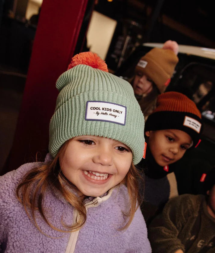 Menina sorridente com gorro Hello Hossy verde-menta com pompom laranja e emblema "COOL KIDS ONLY".
