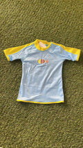 Camisola de verão infantil Btbox, azul claro com mangas amarelas, exibindo um peixe colorido estampado no peito, sobre fundo de relva verde.