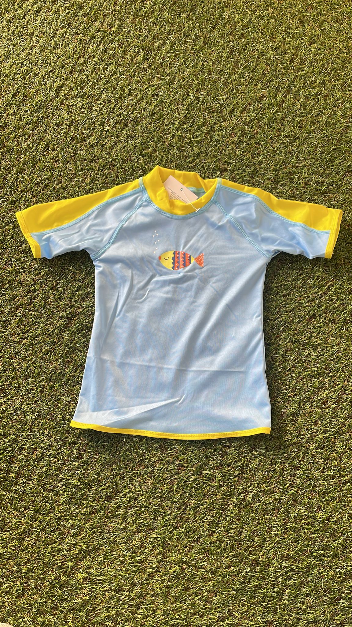 Camisola de verão infantil Btbox, azul claro com mangas amarelas, exibindo um peixe colorido estampado no peito, sobre fundo de relva verde.