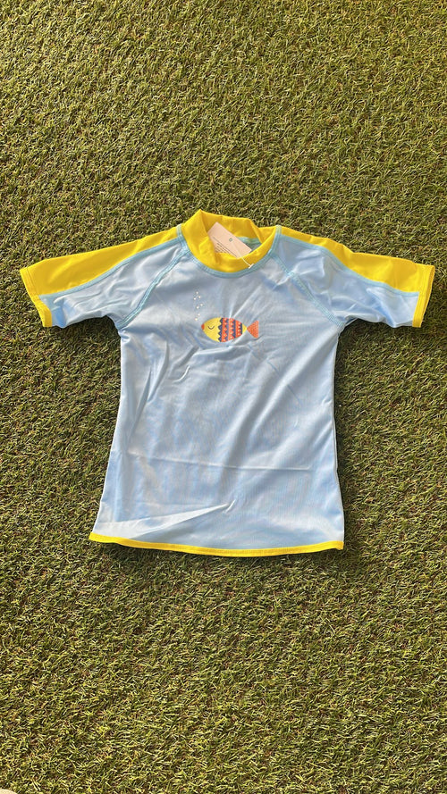 Camisola de verão infantil Btbox, azul claro com mangas amarelas, exibindo um peixe colorido estampado no peito, sobre fundo de relva verde.