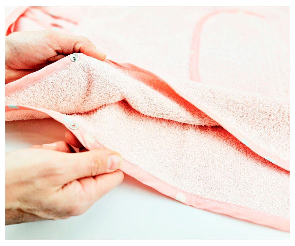 Detalhe das mãos a fechar o fecho de gancho-e-olho lateral de um poncho de banho rosa, mostrando o forro e a textura do algodão.
