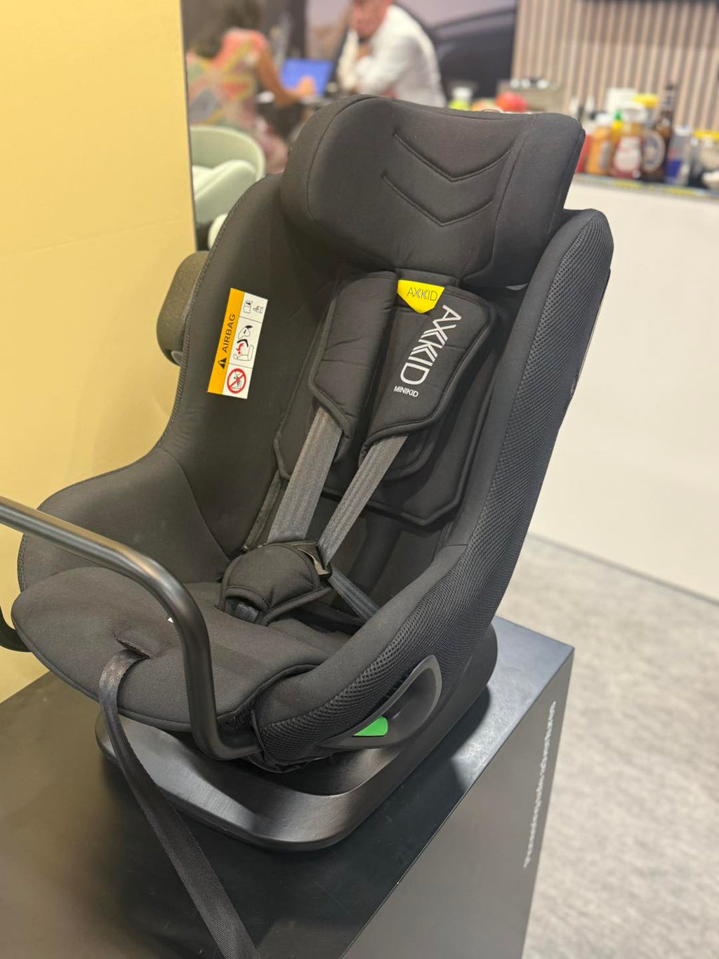 Cadeira auto AXKID Minikid CORE preta em exposição, com etiqueta de aviso de airbag visível e a base de apoio.
