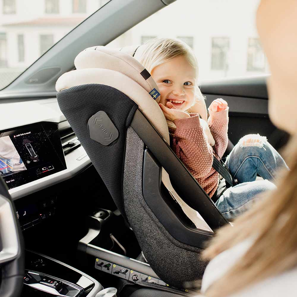 Criança loira sorridente sentada na cadeira auto Axkid ONE 3+ cor bege/creme, instalada em contra marcha num carro, vista pela mãe/pai.