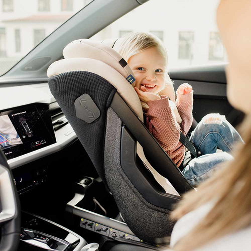 Criança loira sorridente sentada na cadeira auto Axkid ONE 3+ cor bege/creme, instalada em contra marcha num carro, vista pela mãe/pai.