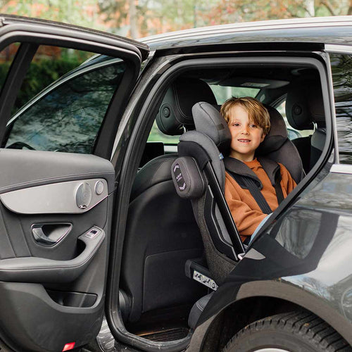 Criança sorridente sentada na Cadeira Auto Axkid ONE 3 instalada contra a marcha num carro, com a porta aberta, destacando a segurança e conforto.