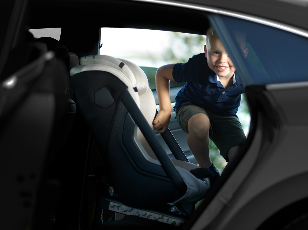 Criança a entrar ou sair da cadeira auto Axkid ONE 3+ cor bege, vista pela porta traseira aberta do carro, mostrando a facilidade de acesso.