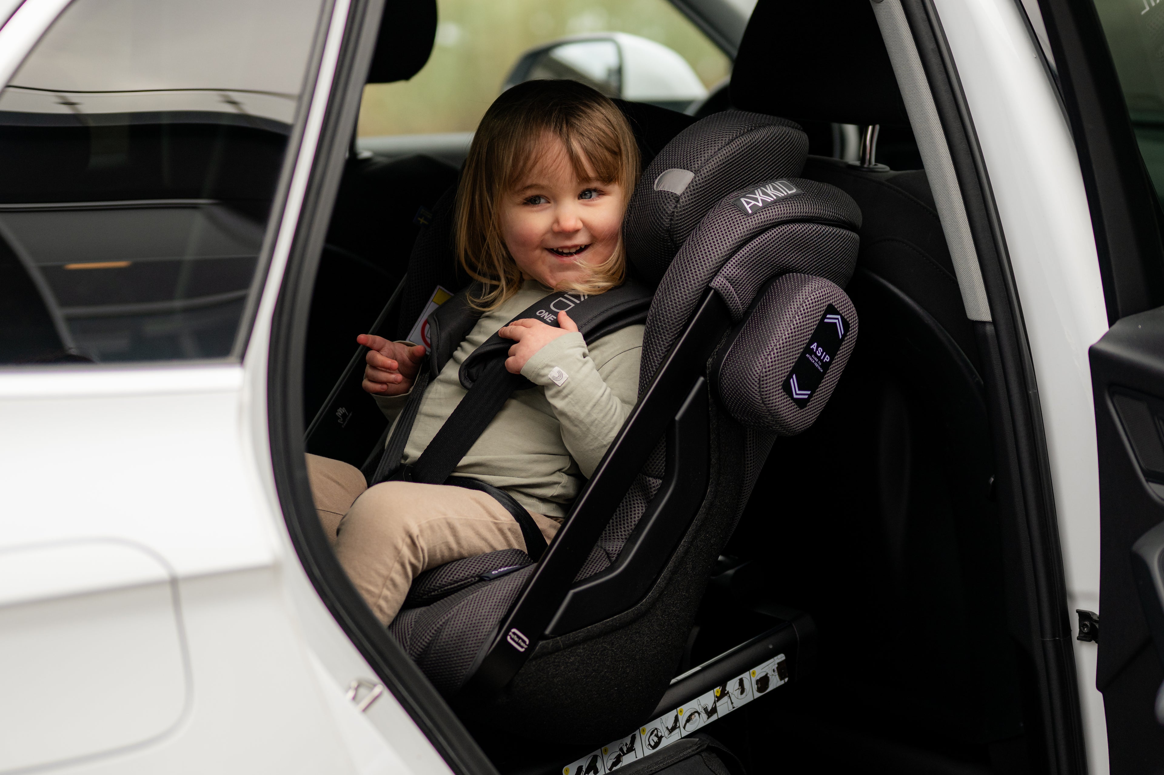 Criança sorridente de cabelo loiro sentada na Cadeira Auto Axkid ONE 3 MESH preta, instalada contra a marcha num carro branco.