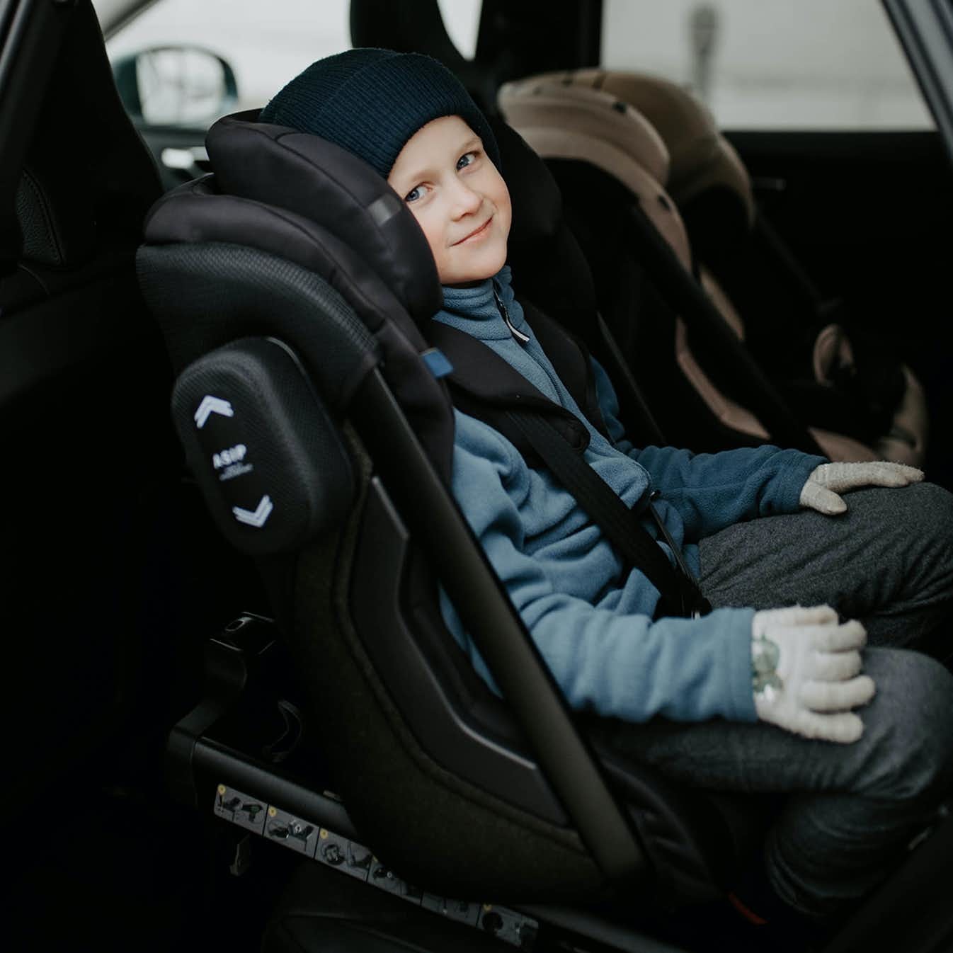 Rapaz mais velho sorridente e confortável na cadeira auto Axkid ONE 3+ preta, instalado em contra marcha, com luvas de inverno.