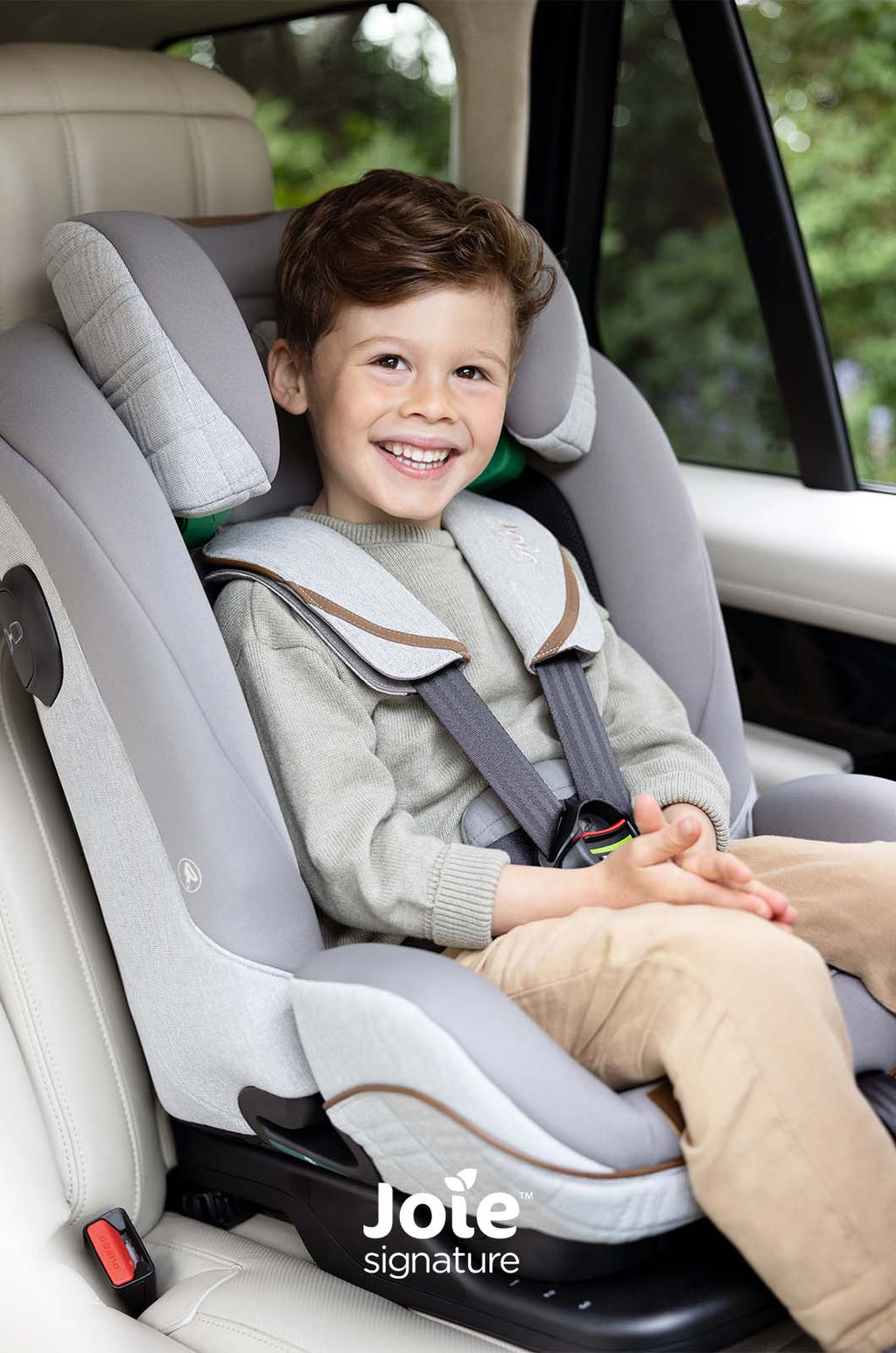 Criança sorridente no assento auto Joie i-Plenti Signature cinzento, com arnês de 5 pontos ajustado, em primeiro plano dentro de um veículo.