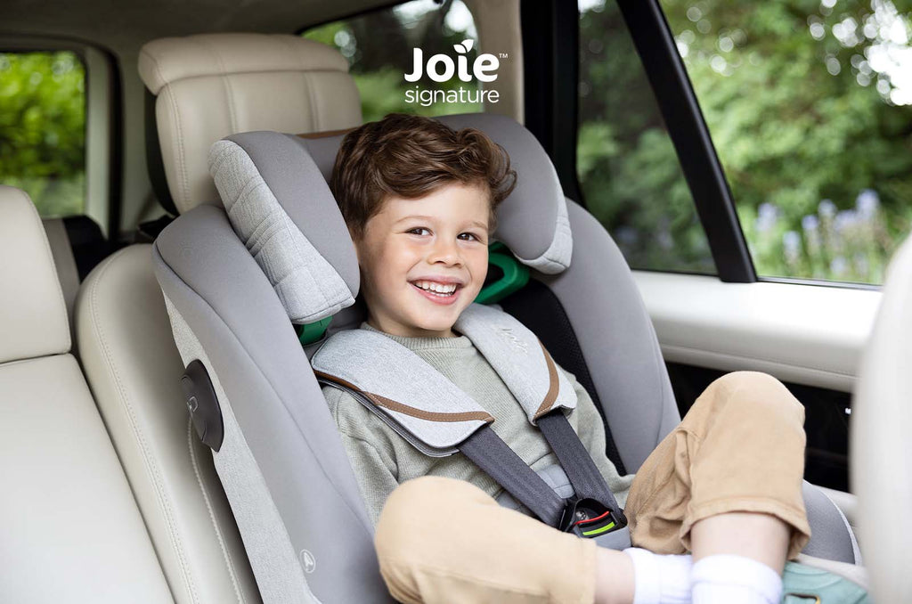Criança sorridente no assento auto Joie i-Plenti Signature cinzento, com foco no conforto e ajuste do encosto de cabeça, dentro de um carro.