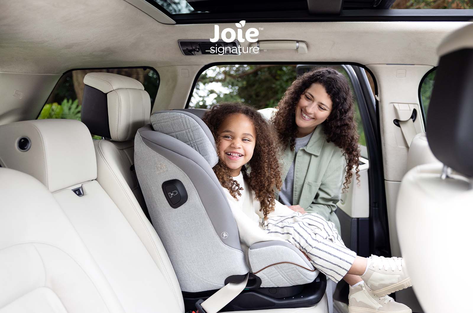 Criança sorridente no assento auto Joie i-Plenti Signature cinzento claro, instalada num carro com bancos de pele clara, com a mãe ao lado.