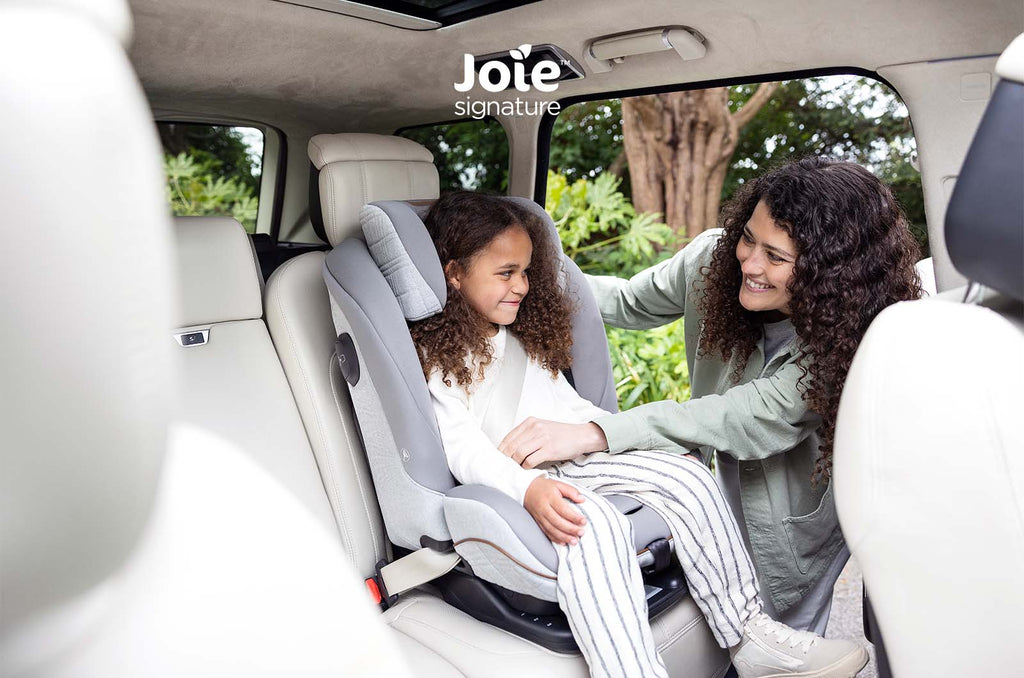 Criança sorridente sentada na cadeira auto Joie i-Plenti Signature cinzenta, instalada num carro com interiores claros, enquanto uma mulher a ajuda a ajustar o arnês.