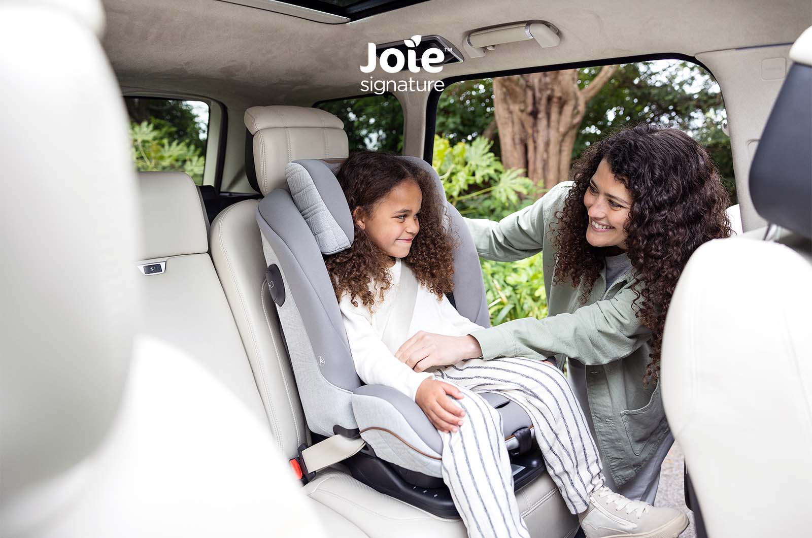 Criança sorridente sentada na cadeira auto Joie i-Plenti Signature cinzenta, instalada num carro com interiores claros, enquanto uma mulher a ajuda a ajustar o arnês.