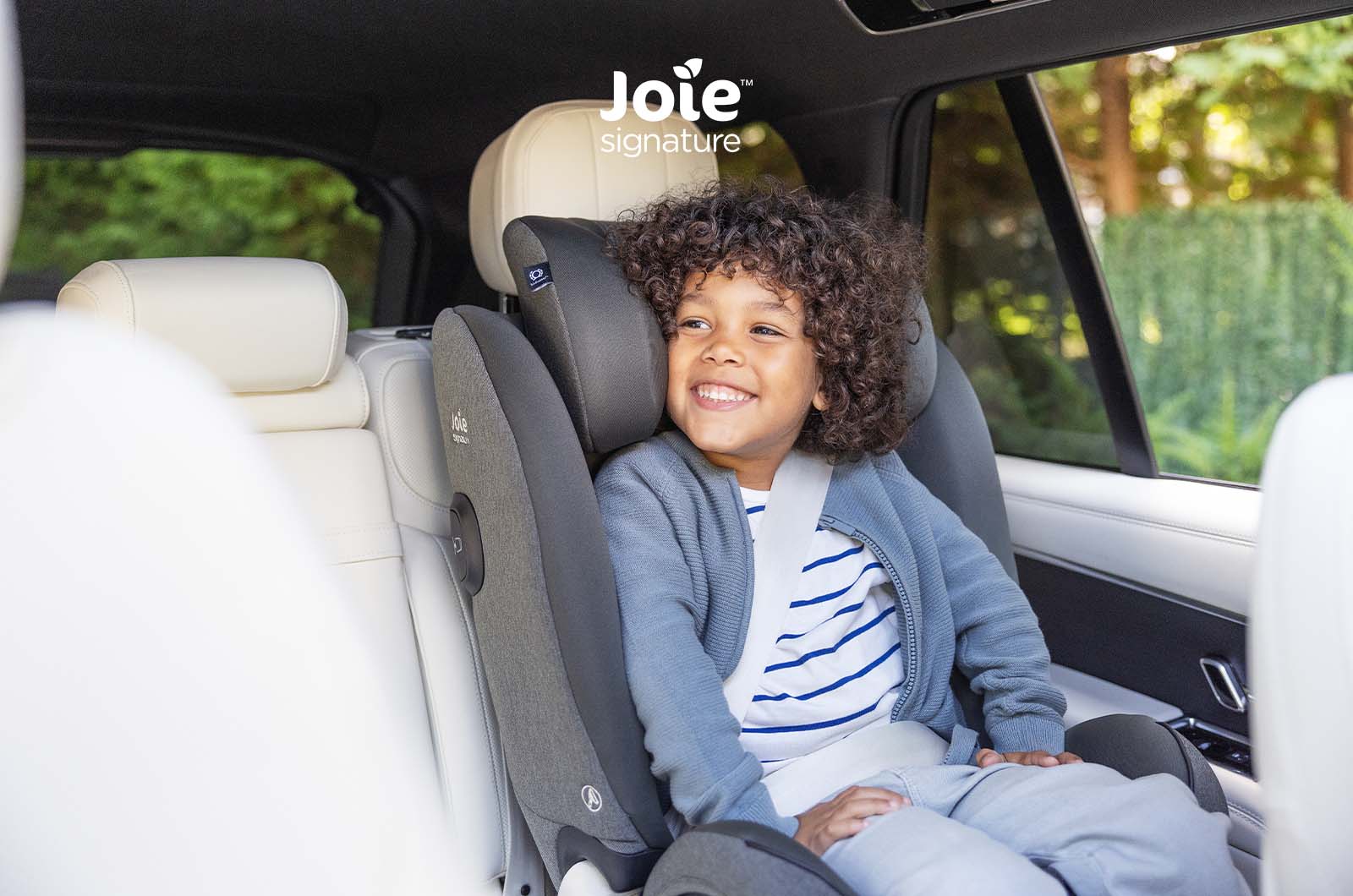 Criança sorridente com cabelo encaracolado sentada na cadeira auto Joie i-Spin XL Signature cinzenta, instalada num carro com interiores de pele clara.