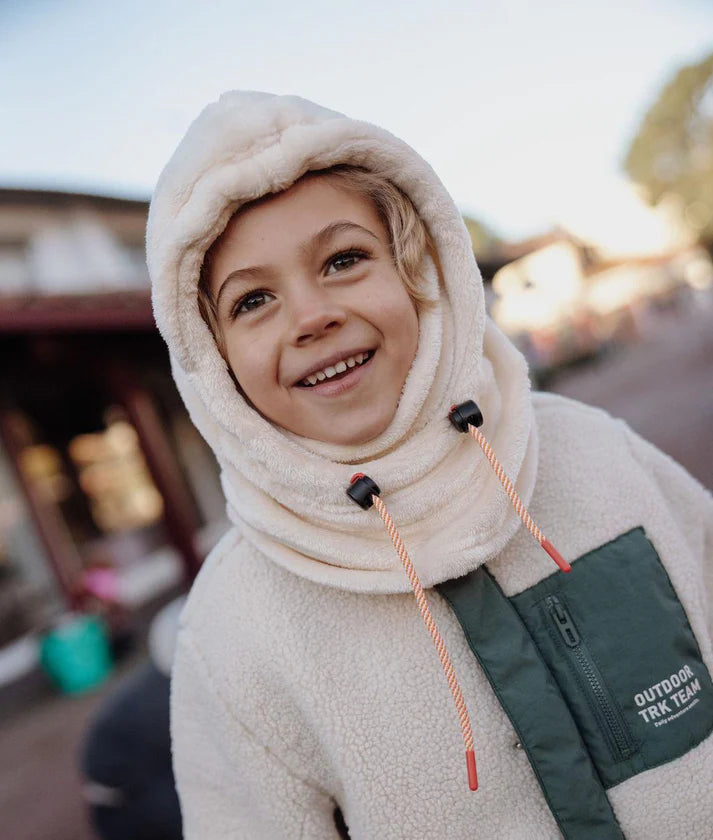 Criança sorridente a usar o gorro Hello Hossy Warmy Cream com capuz e atilhos ajustáveis, em ambiente exterior.