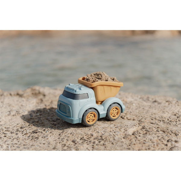 Camião basculante Little Dutch azul com areia na caçamba amarela, pousado numa pedra junto à água.