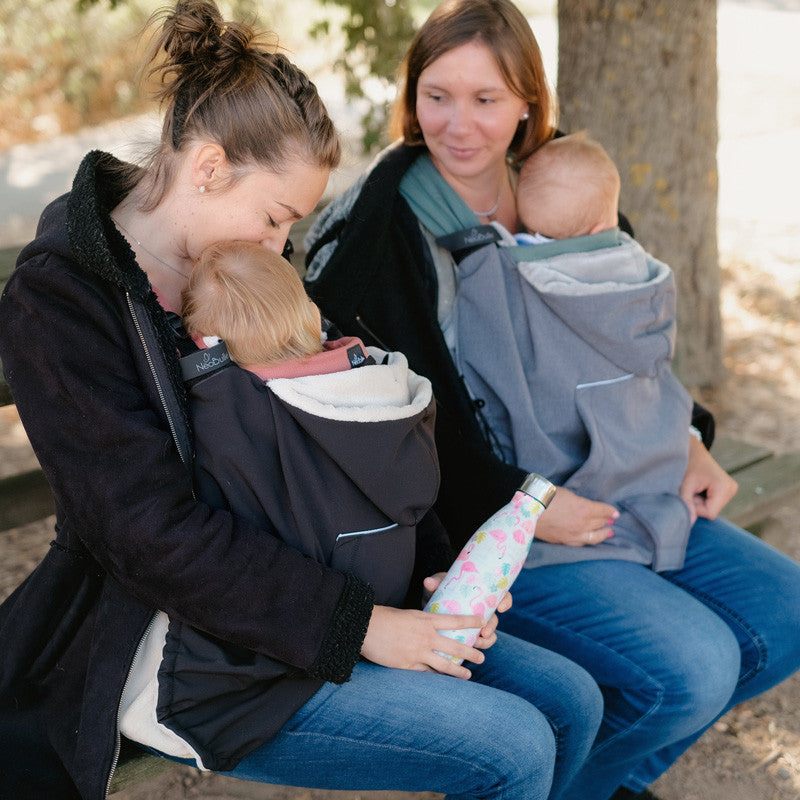 Duas mães sentadas num banco, ambas a usar capas de babywearing (uma preta e uma cinzenta), demonstrando a versatilidade do acessório.