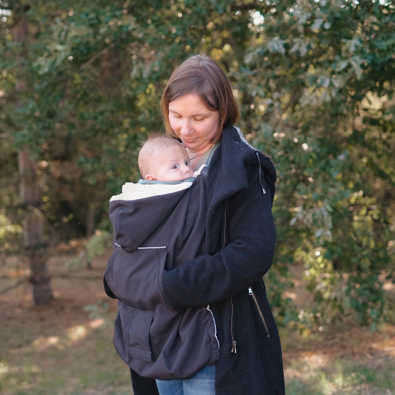 Mulher a usar a capa de babywearing Neobulle preta, com o bebé aconchegado, em ambiente exterior com árvores.