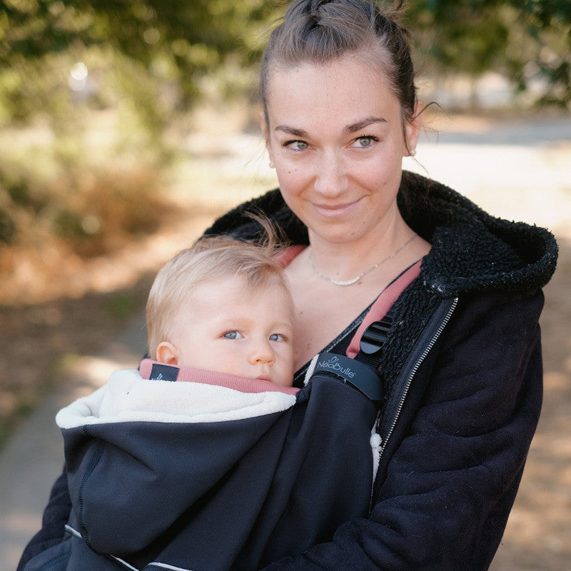 Mãe sorridente a usar a capa de babywearing Neobulle preta com o seu bebé, mostrando o ajuste confortável e o forro claro.