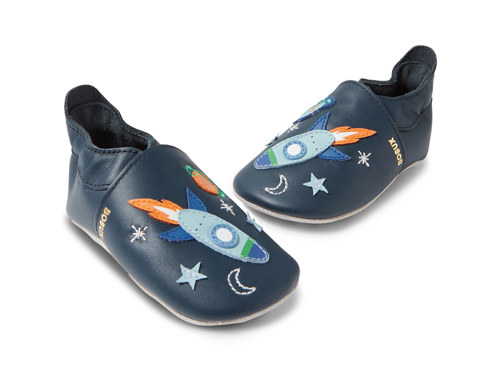 Par de sapatos Bobux Cosmic Rocket Soft Sole em azul-marinho, vistos de lado em ângulo, com detalhes de foguetões e estrelas.