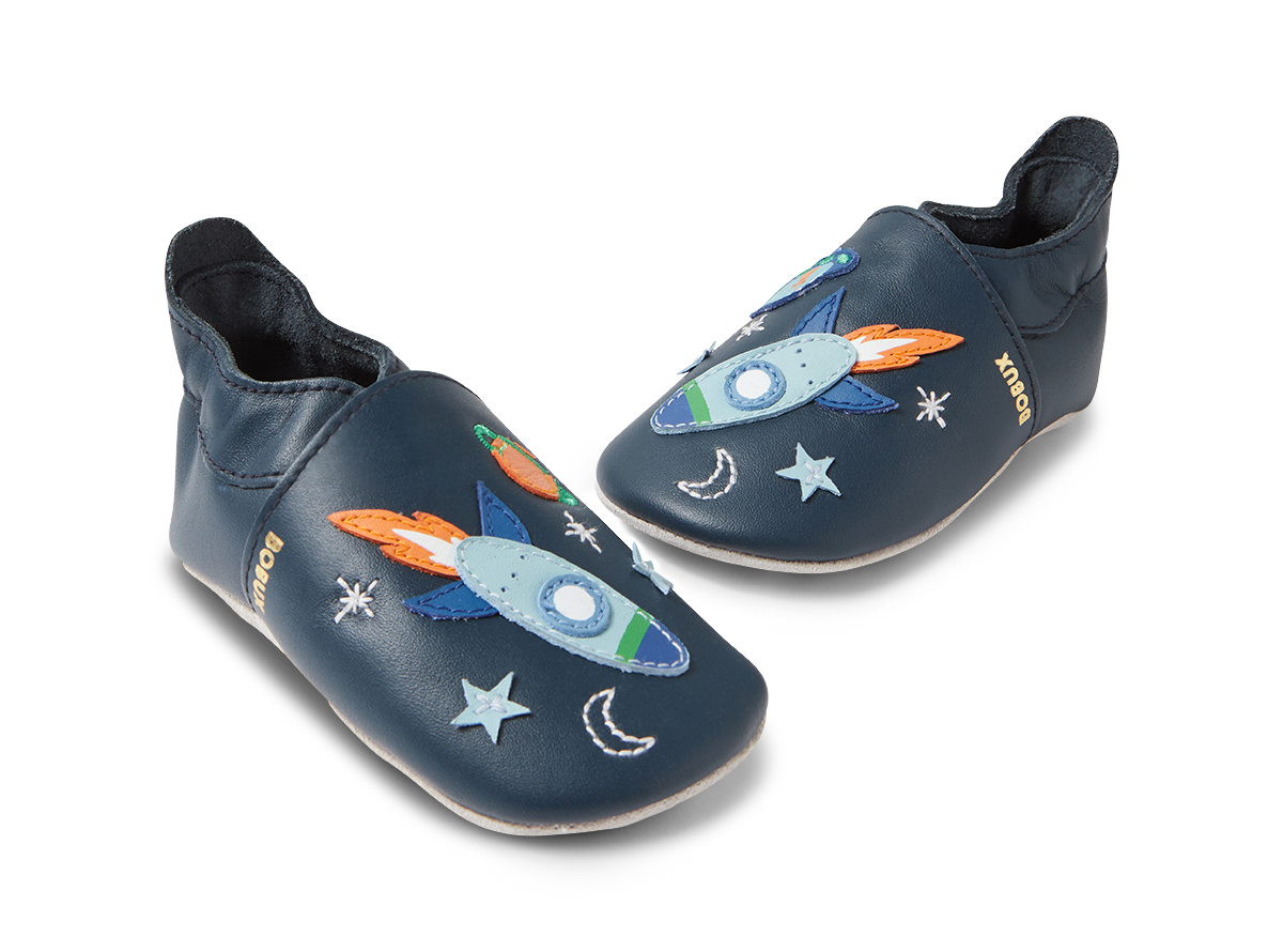 Par de sapatos Bobux Cosmic Rocket Soft Sole em azul-marinho, vistos de lado em ângulo, com detalhes de foguetões e estrelas.