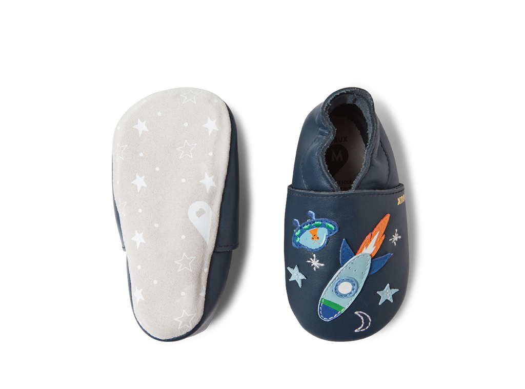 Um sapato Bobux Cosmic Rocket Soft Sole em azul-marinho, visto de cima, e a sola macia do outro sapato com padrão de estrelas.