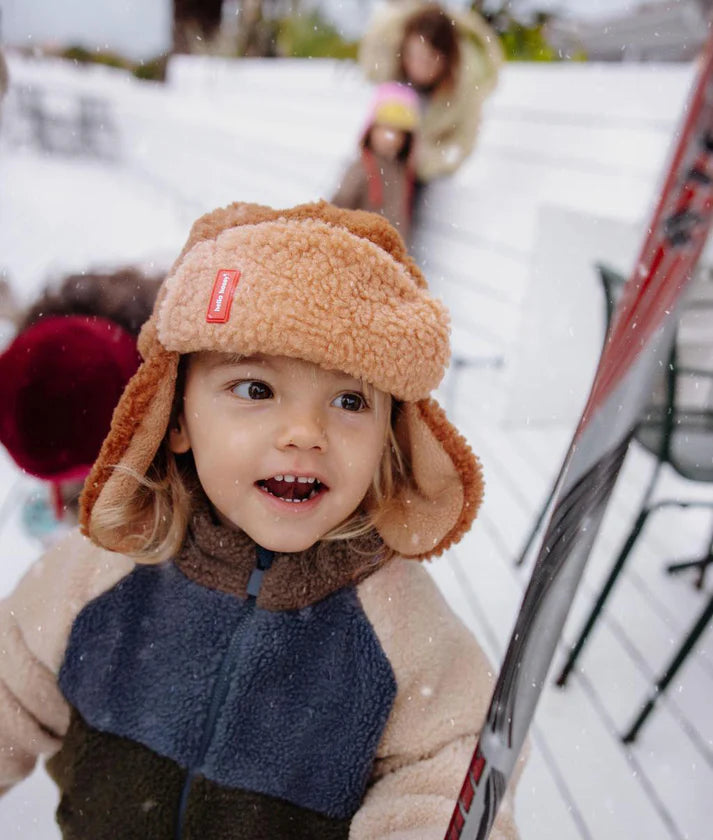 Criança com gorro aviador Hello Hossy Cozy Sven castanho claro e casaco polar multicolorido, com esqui ao lado, a brincar na neve.