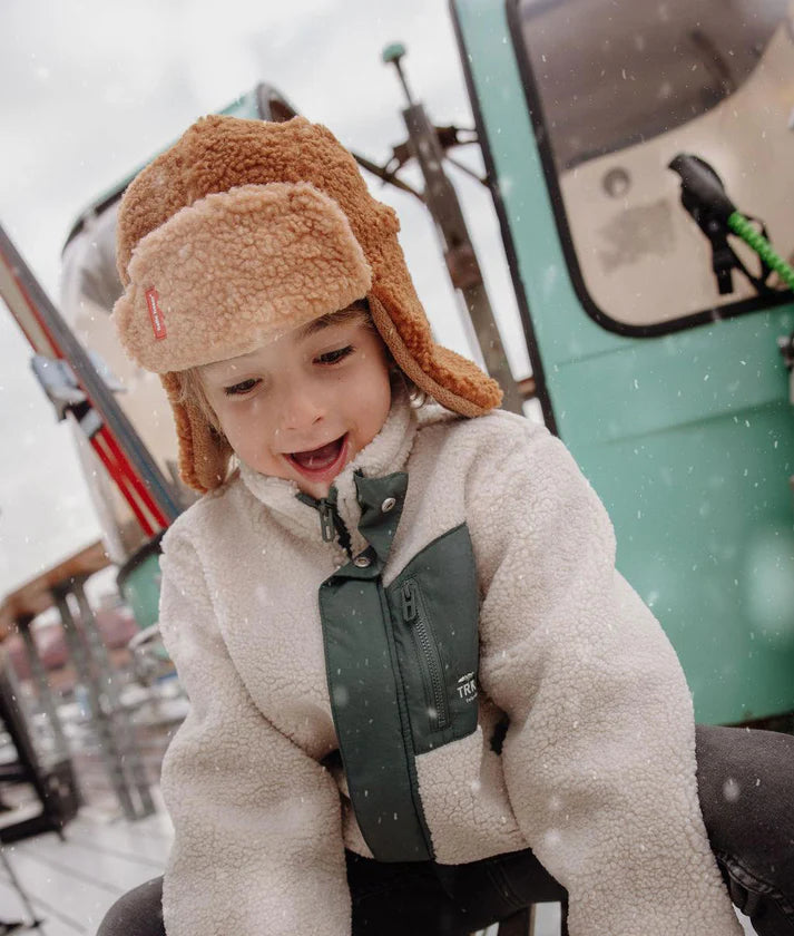 Criança sorridente com gorro aviador Hello Hossy Cozy Sven castanho claro e casaco polar branco e verde, a brincar na neve.