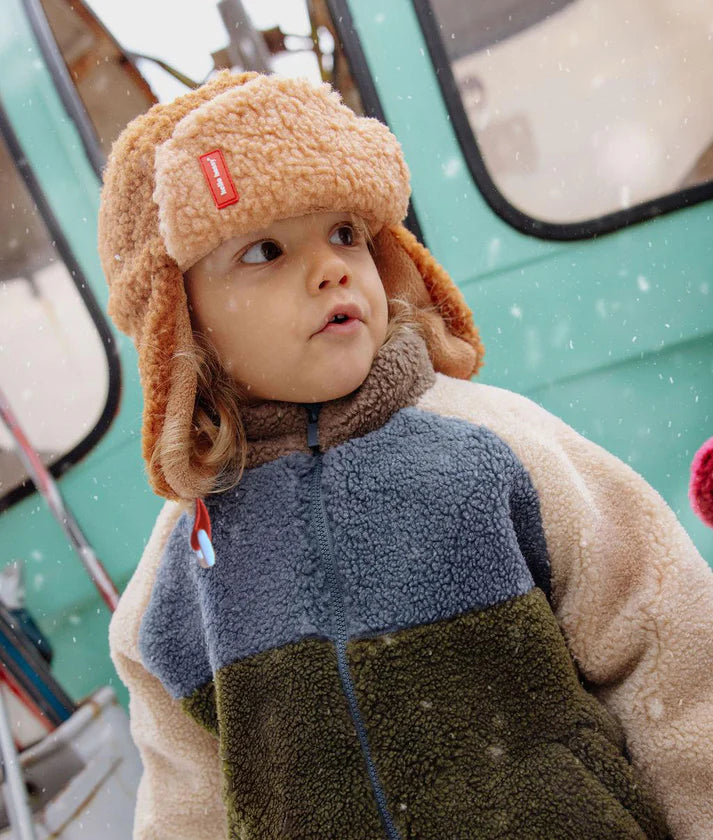 Criança com gorro aviador Hello Hossy Cozy Sven castanho claro e casaco polar multicolorido, a olhar para cima, em ambiente nevado.