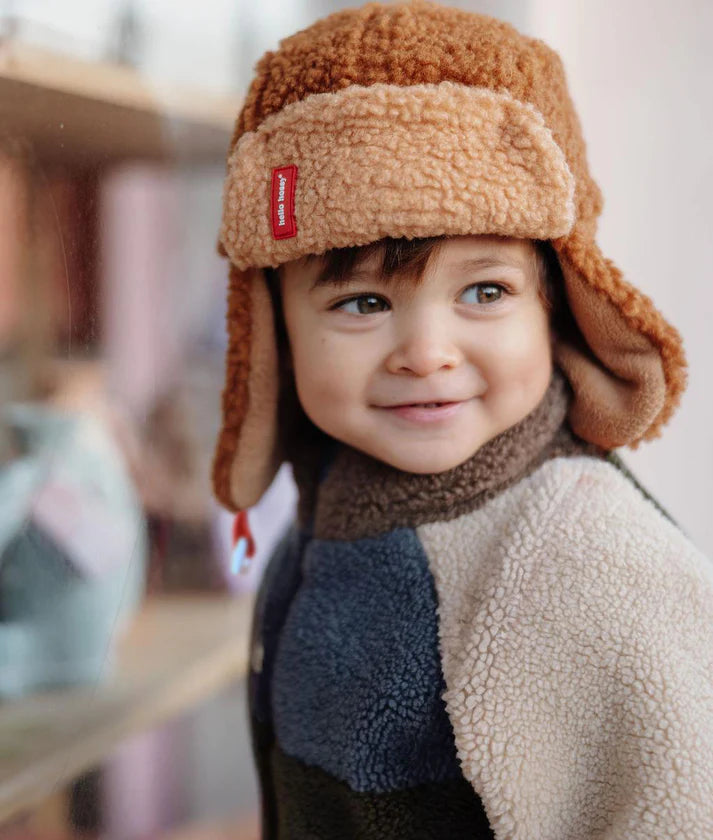 Menino com gorro aviador Hello Hossy Cozy Sven castanho claro, sorridente, com casaco polar multicolorido, a olhar para o lado.