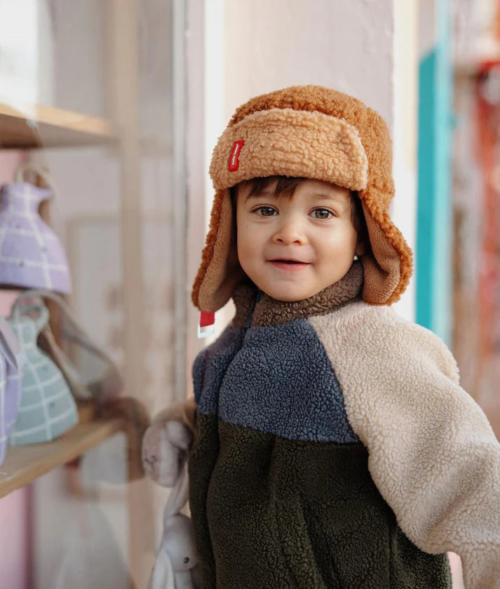 Menino com gorro aviador Hello Hossy Cozy Sven castanho claro, a sorrir para a câmara, com casaco polar multicolorido, em interior.