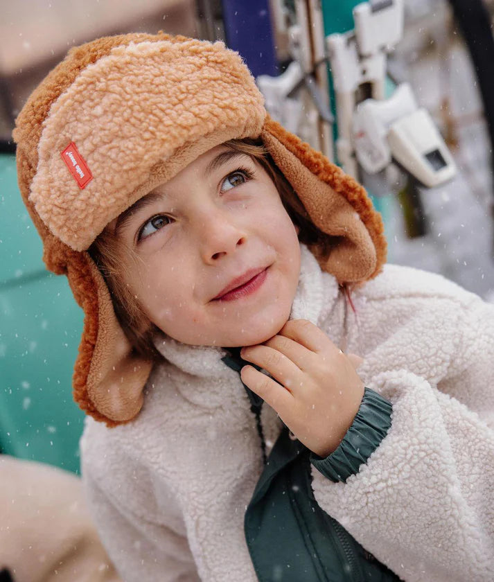 Criança com gorro aviador Hello Hossy Cozy Sven castanho claro e casaco polar branco, a tocar no queixo, a olhar para cima na neve.