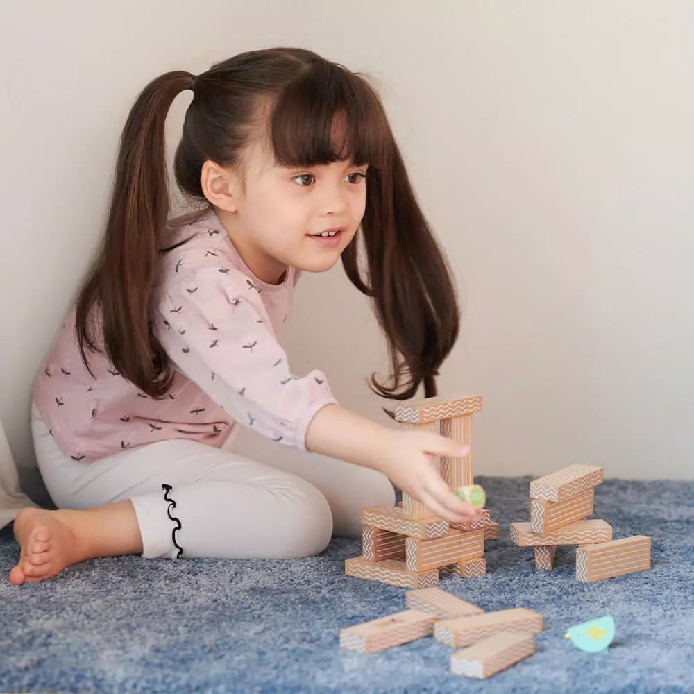 Menina sorridente a interagir com as peças do jogo de empilhar pássaros, estendendo a mão para um bloco no chão.
