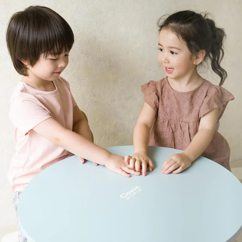 Duas crianças sorridentes interagem junto à Mesa de Madeira Grace da Classic World, com tampo redondo azul-água.