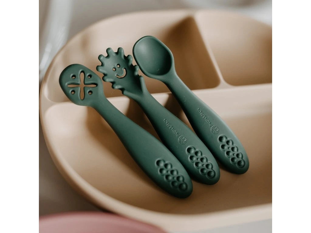 Três colheres de silicone verde-escura alinhadas sobre um prato de alimentação bege com divisórias, mostrando detalhes das pontas e texturas da pega.