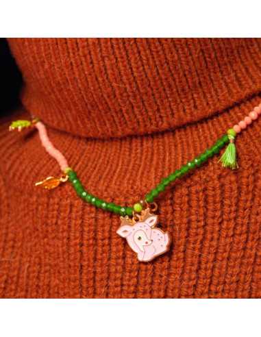 Close-up de um colar infantil com contas cor-de-rosa e verde, apresentando um pendente de biche cor-de-rosa e um pompom verde, usado sobre um camisola de gola alta cor de tijolo.