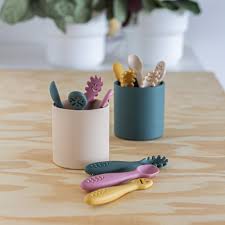 Conjunto de três colheres de silicone coloridas (verde-escura, rosa, amarela e bege) dispostas em dois recipientes cilíndricos e três espalhadas sobre uma superfície de madeira clara.