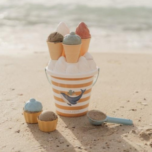 Conjunto de gelados de areia Little Dutch pousado na areia da praia, com o mar ao fundo, mostrando o balde, a colher e alguns gelados já moldados.