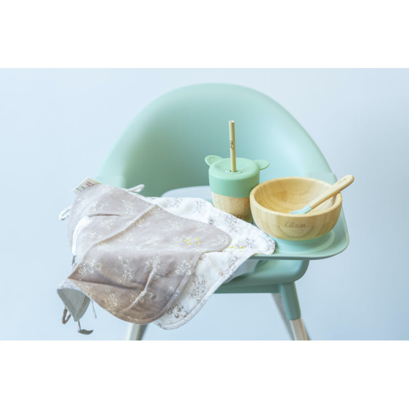 Conjunto de artigos de refeição Citron, incluindo um babete de tecido, taça de bambu e copo de bambu com tampa de urso, dispostos numa cadeira alta de bebé verde.
