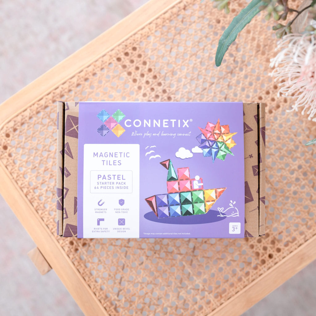 Caixa do Connetix Jogo Blocos Magnético Pastel Starter 64 pc pousada numa cadeira de verga com folhagem ao fundo.