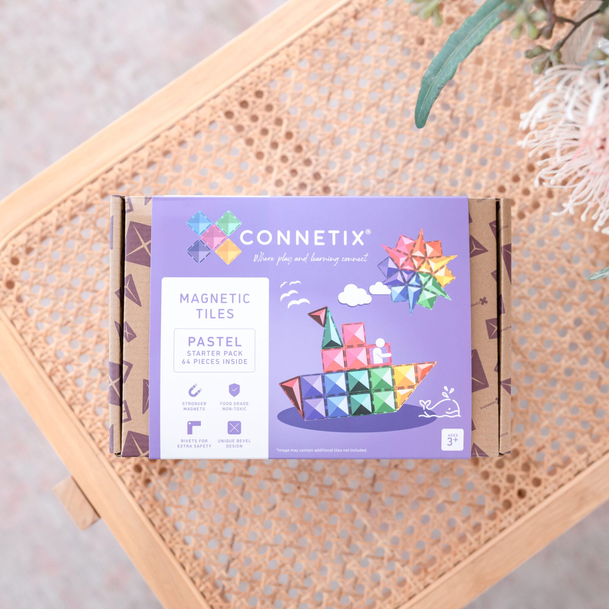 Caixa do Connetix Jogo Blocos Magnético Pastel Starter 64 pc pousada numa cadeira de verga com folhagem ao fundo.