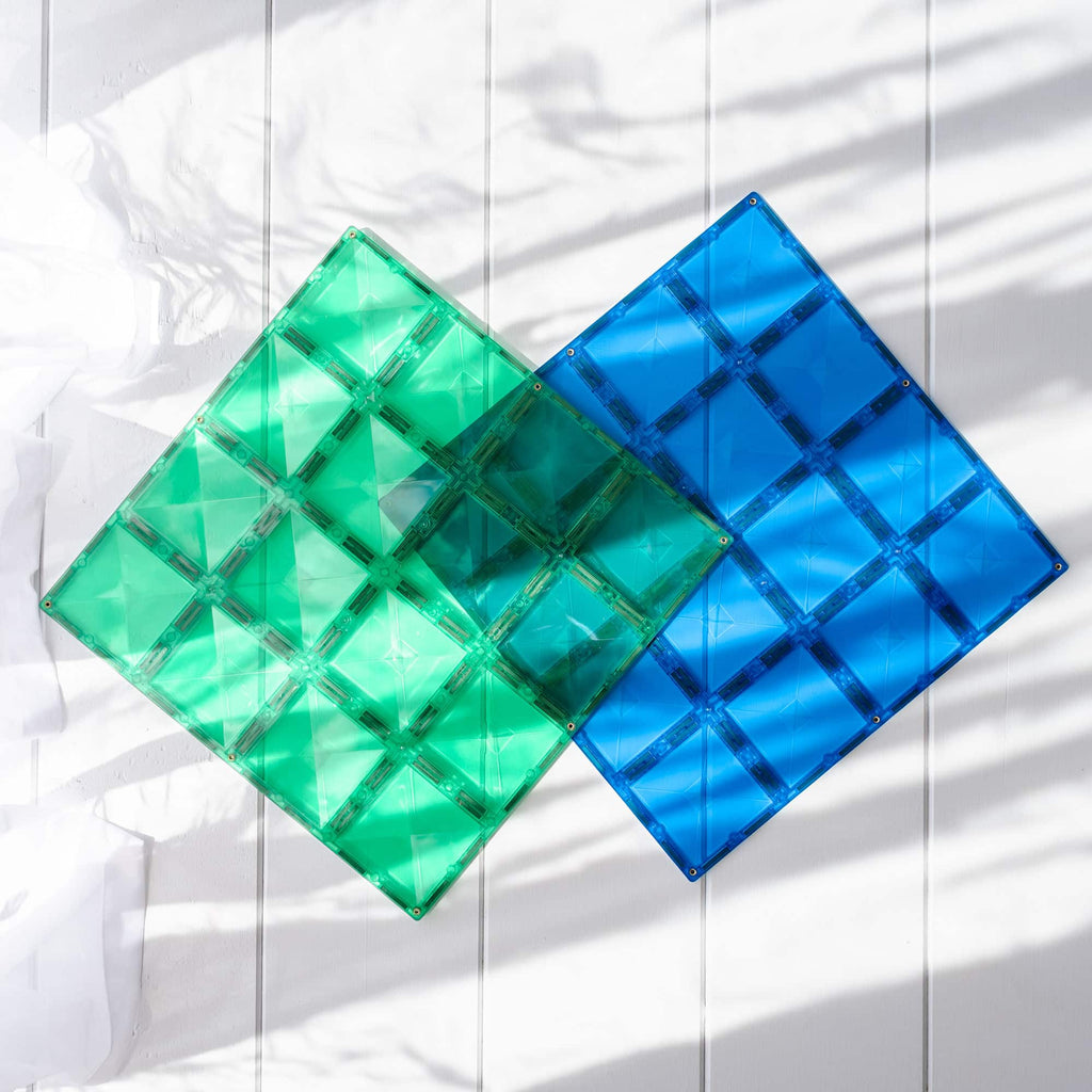 Duas placas base magnéticas Connetix quadradas, uma verde esmeralda e outra azul, sobre superfície branca com sombras de folhas.