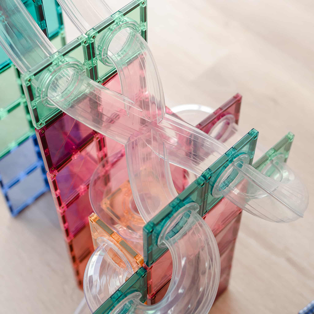 Detalhe de tubos transparentes e peças magnéticas Connetix em tons pastel (rosa, verde) a formar uma rampa de bolas complexa.