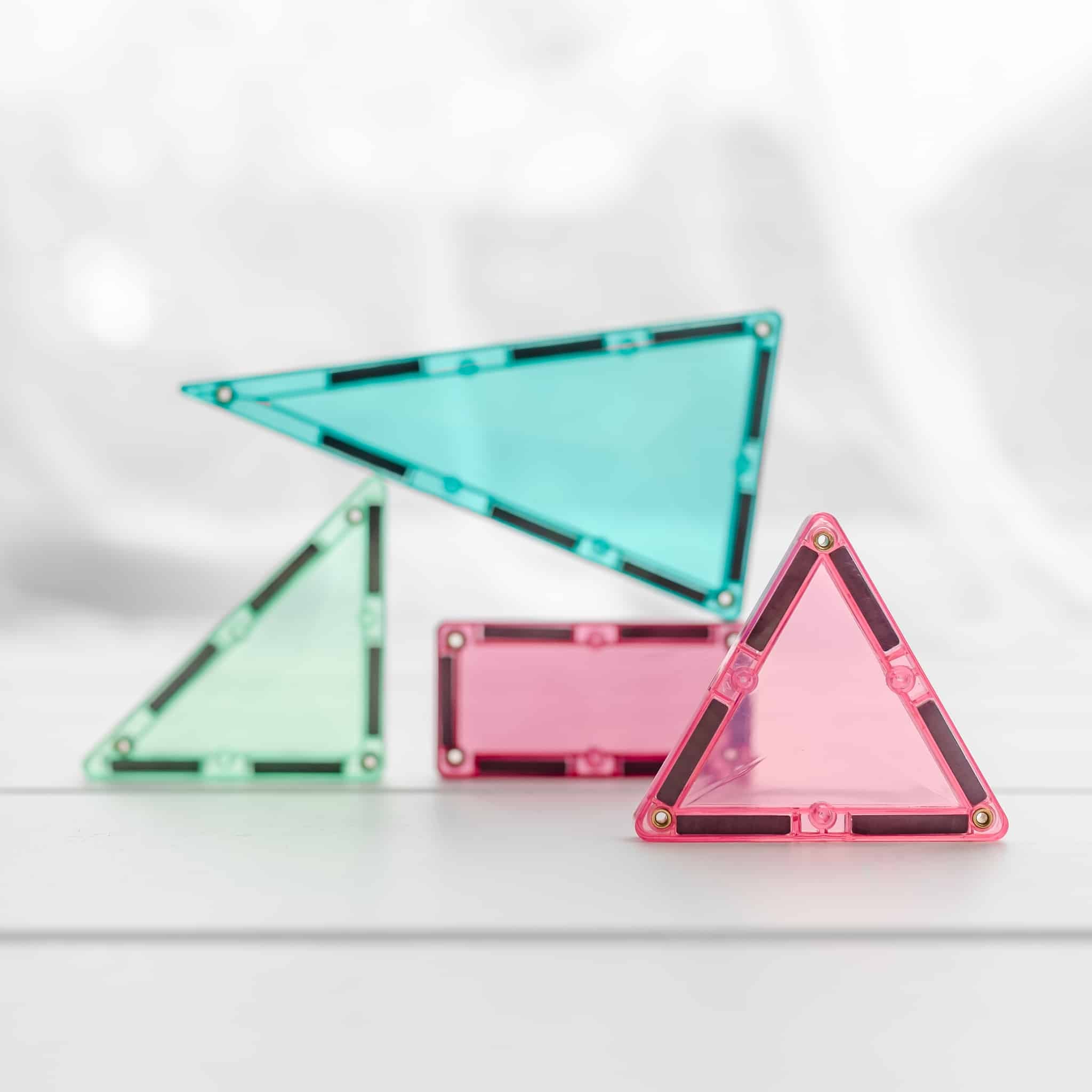 Quatro peças magnéticas Connetix em cores pastel (rosa, verde, azul-turquesa) em primeiro plano, demonstrando a transparência e as formas de construção.