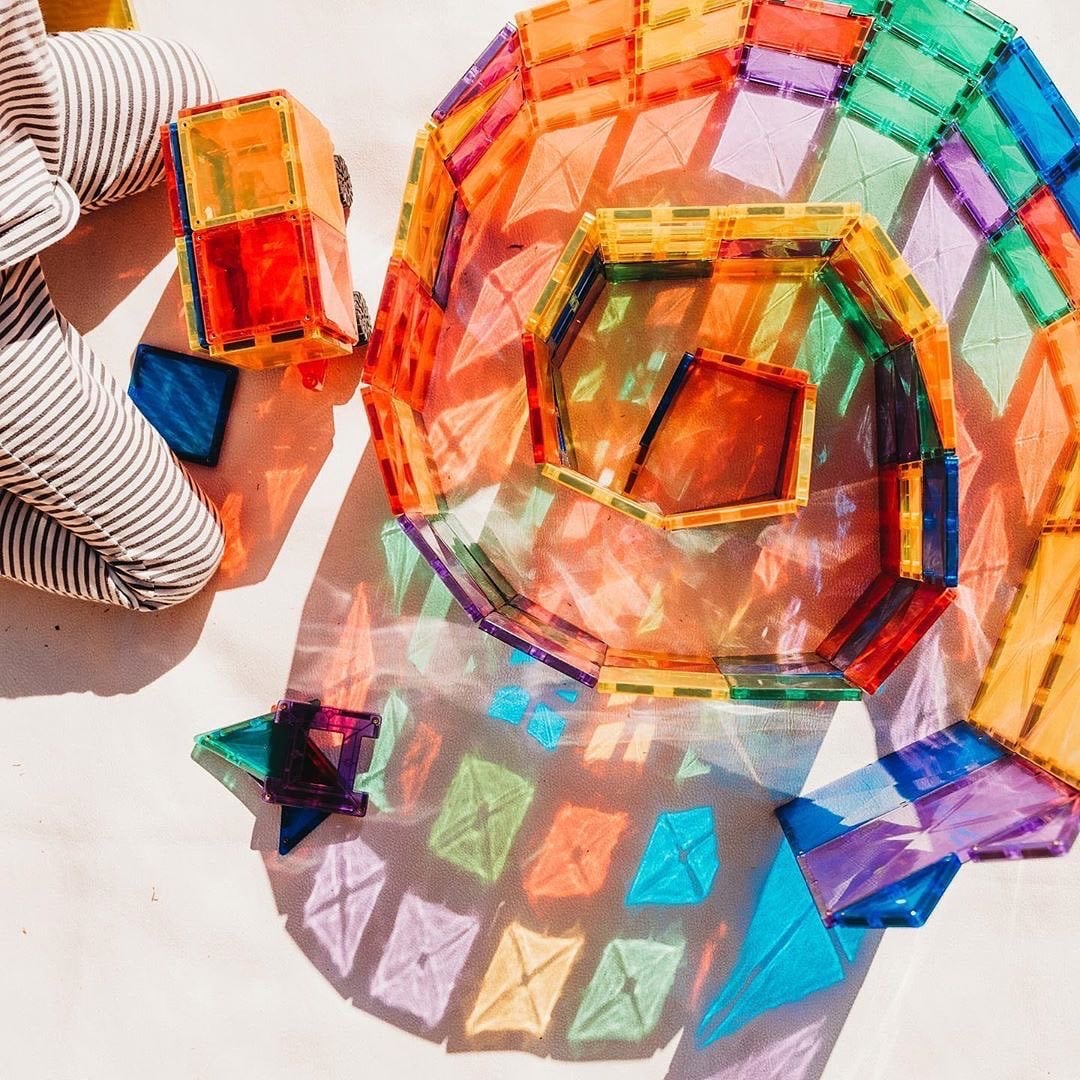Mãos a construir espiral com blocos magnéticos Connetix Rainbow, mostrando refrações de luz coloridas.