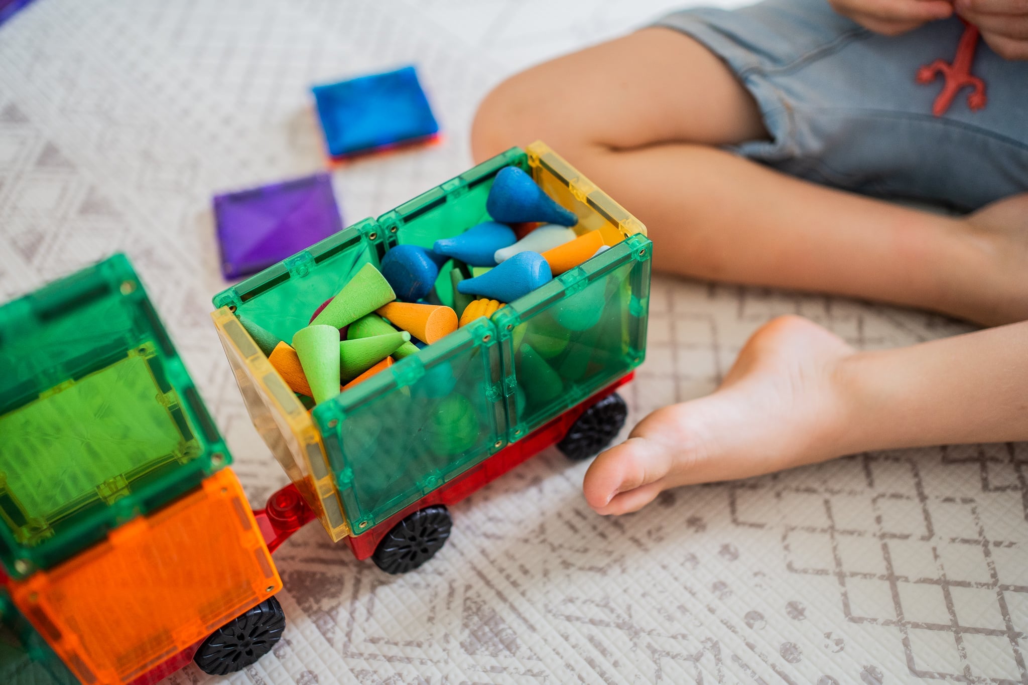 Criança a brincar no chão com um camião construído com blocos magnéticos Connetix Motion, carregado com peças de madeira coloridas.