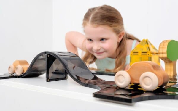 Criança sorridente a interagir com uma rampa de estrada magnética preta Connetix e carros de madeira, focada na brincadeira.