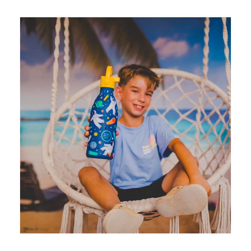 Criança sorridente sentada numa cadeira de baloiço na praia, segurando a garrafa térmica Cool Bottles Galactic Ride 500ml com design espacial.